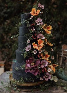 a black cake with flowers on it sitting on top of a stone slab in front of some bushes