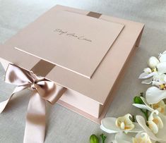 a pink box with a ribbon and flowers on the table next to it is a bouquet of white orchids