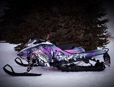 a purple and black snowmobile parked in the snow next to a large pine tree