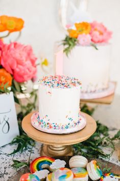 two birthday cakes with sprinkles are on a table next to cookies and flowers