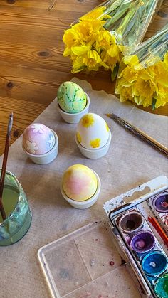 an assortment of painted eggs sitting on a table with flowers and paintbrushes in front of them