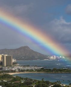 a rainbow in the sky over a city by the ocean with buildings and mountains behind it