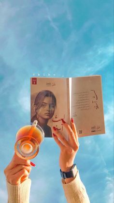 a person holding up a book and a drink in front of a blue sky with clouds