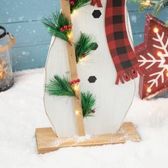 a snowman made out of wood and decorated with christmas lights in front of a snowy background