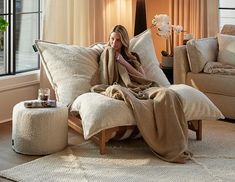 a woman wrapped in a blanket is sitting on a chair with pillows and a footstool