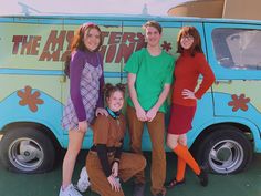 four people are posing in front of a van with the words the mystery machine on it