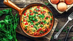 an omelet in a pan with vegetables and eggs on a wooden table next to utensils