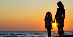 two people standing on the beach holding hands as the sun sets in the distance behind them