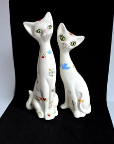 two white ceramic cats sitting next to each other on a black background, one has green eyes and the other has an orange nose