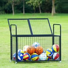 a metal basket filled with lots of balls on top of a grass covered field in front of trees