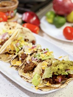 three tacos are sitting on a white plate next to some tomatoes and other vegetables