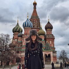 a woman standing in front of a building with domes on it's sides and wearing a fur hat