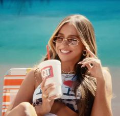 a beautiful woman sitting on top of a beach chair holding a cup and wearing glasses