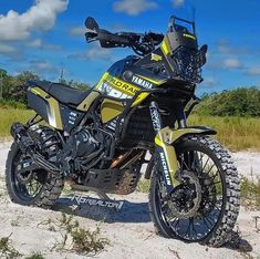 a yellow and black motorcycle parked on top of a sandy beach next to trees in the background