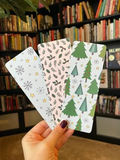 someone holding up three christmas cards in front of a bookshelf