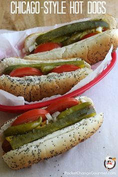 two hot dogs in buns with pickles and tomatoes on them, sitting next to each other