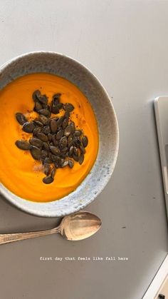 a bowl of soup with pumpkin seeds in it next to a spoon and laptop computer