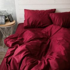 a bed with red sheets and pillows in a room next to a small side table