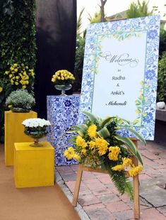 a welcome sign with yellow flowers and greenery in front of it on a brick walkway