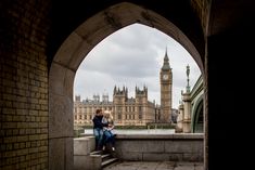 a man and woman are sitting on a ledge in front of the big ben clock tower