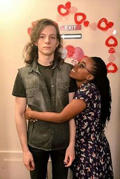 two people standing next to each other in front of a wall with hearts on it