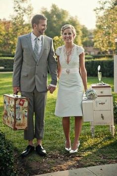 a man and woman standing in the grass holding hands with suitcases on their feet