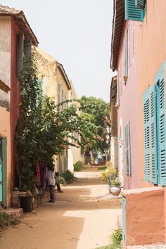 two people are walking down an alley way with blue shutters on each side and pink buildings in the background