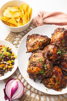 some chicken wings are on a white plate next to a bowl of beans, corn and tortilla chips