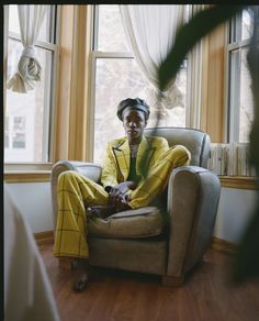 a woman sitting in a chair with her legs crossed and wearing a yellow suit, looking out the window