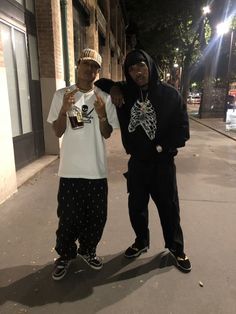 two young men standing next to each other on a sidewalk at night with their arms around one another