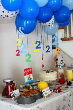 a table topped with blue balloons and cake next to a white table cloth covered table