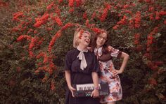 two young women standing next to each other in front of red flowers and trees with their hands up