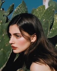 a woman with long hair standing next to a green plant and looking off into the distance