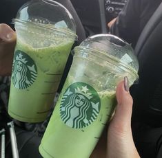 two people holding up starbucks cups with green liquid on them in the backseat of a car