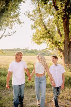 three people holding hands walking down a dirt path in front of a tree and grass field