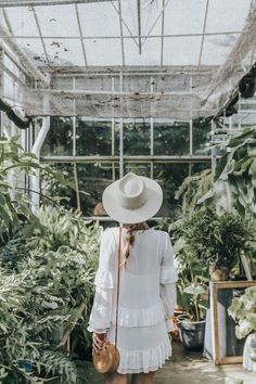 a woman in a white dress and hat walking through a greenhouse filled with potted plants