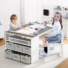 two children are sitting at a table with paint and pencils on it, while another child is standing in the background