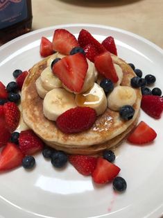 pancakes with bananas, strawberries and blueberries on a white plate next to a bottle of booze