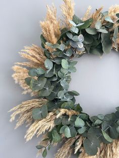 a wreath made out of dried grass and eucalyptus leaves on a white wall with grey background