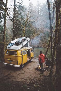 a yellow van parked in the woods next to a man with a surfboard on top