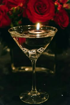 a candle is lit in a martini glass with dice on the rim and red roses behind it