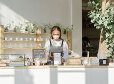 a woman wearing a face mask behind a counter at a cafe with plants on the shelves