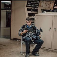 a man sitting in a chair holding a camera and looking at his cell phone while he sits next to a locker