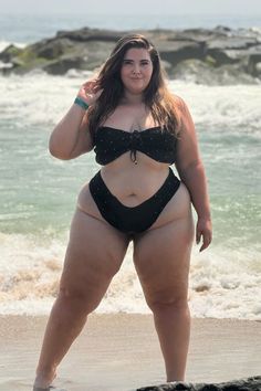 A woman stands confidently at the edge of a sandy beach, wearing a black bikini with subtle sparkles. Waves crash behind her, with large rocks in the background