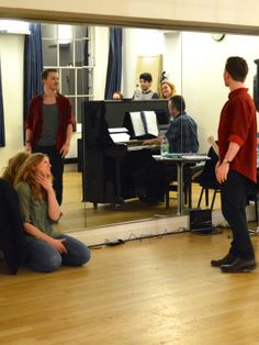 a group of young men and women standing around a living room with piano in the background