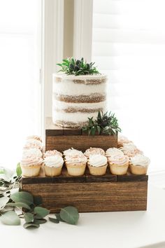 a three tiered cake and cupcakes on a wooden stand with greenery