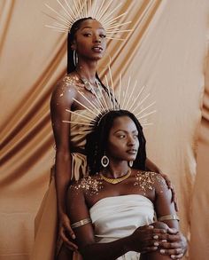 two women in white dresses with gold decorations on their heads and arms, one woman is sitting down while the other stands behind her