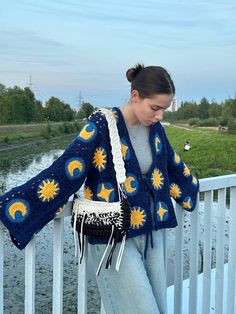 a woman standing on a bridge wearing a blue and yellow crochet sweater with sunflowers