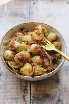 a bowl filled with potatoes and onions on top of a wooden table next to a fork