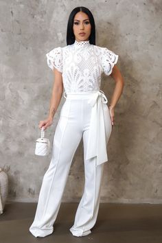 a woman standing in front of a wall wearing white pants and a top with lace on it